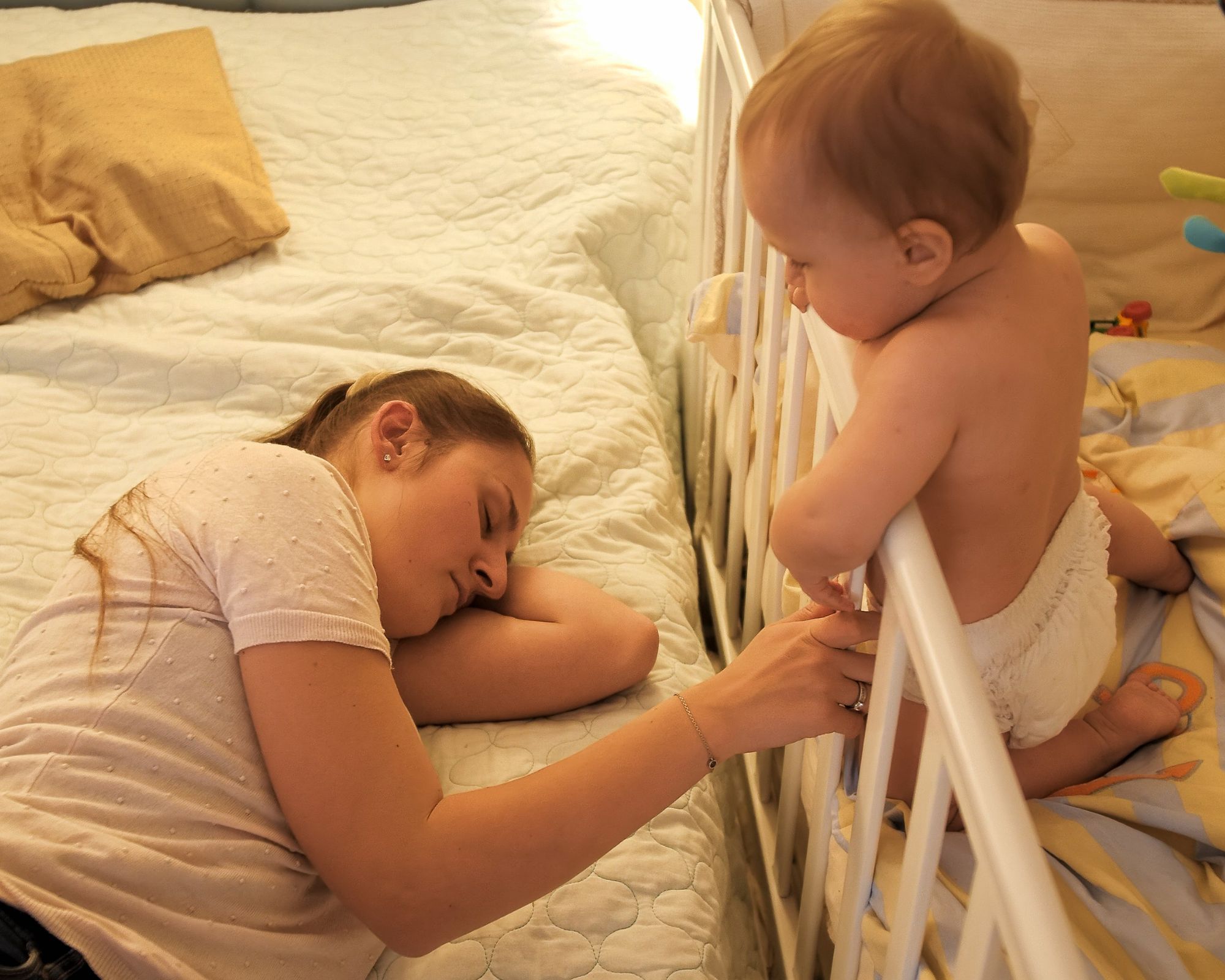 Mutter schläft neben dem Babybett, indem das Baby wach steht 